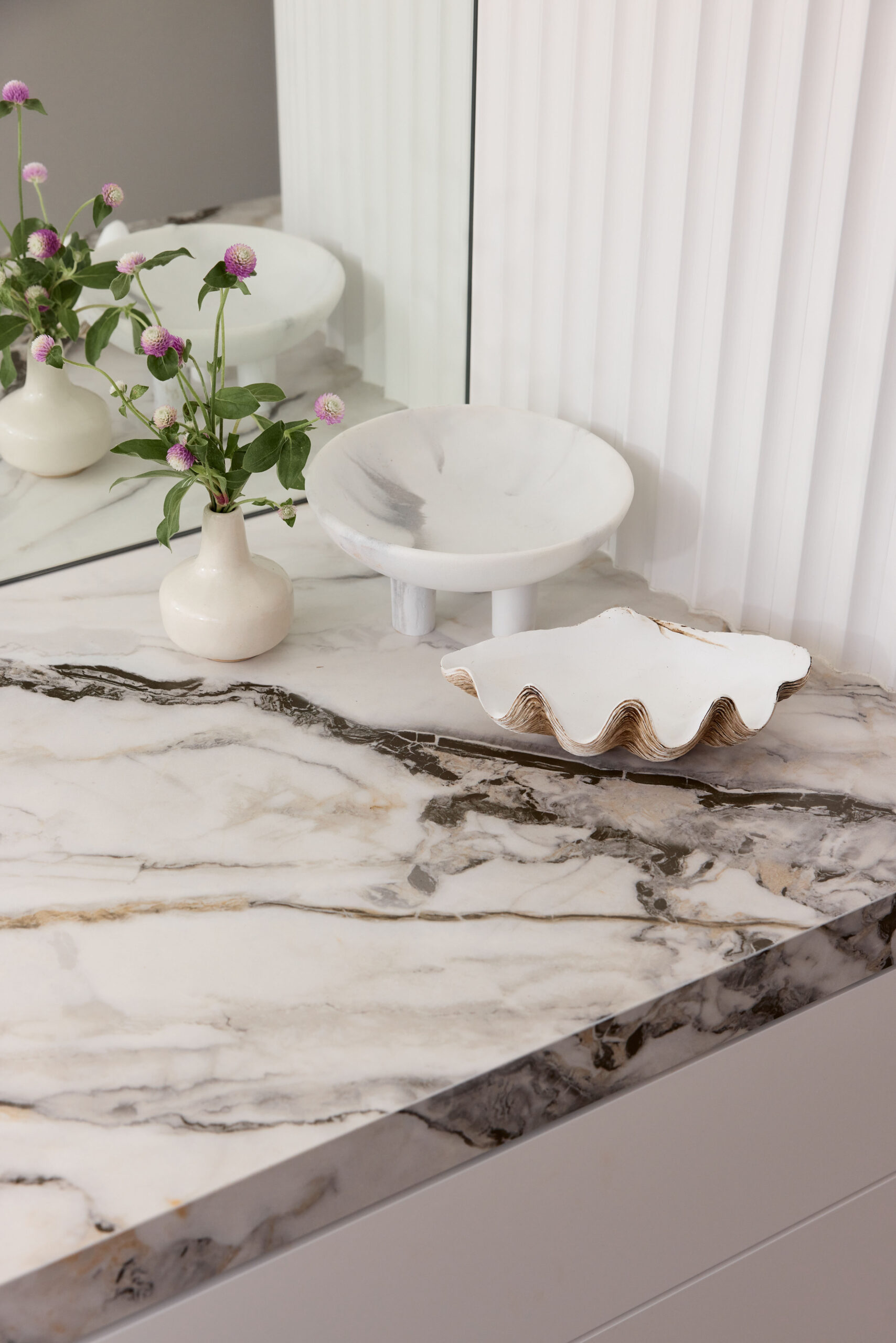 White ceramic vase with pink globe flowers on a marble countertop beside a round white bowl and a scalloped shell tray near a mirror.
