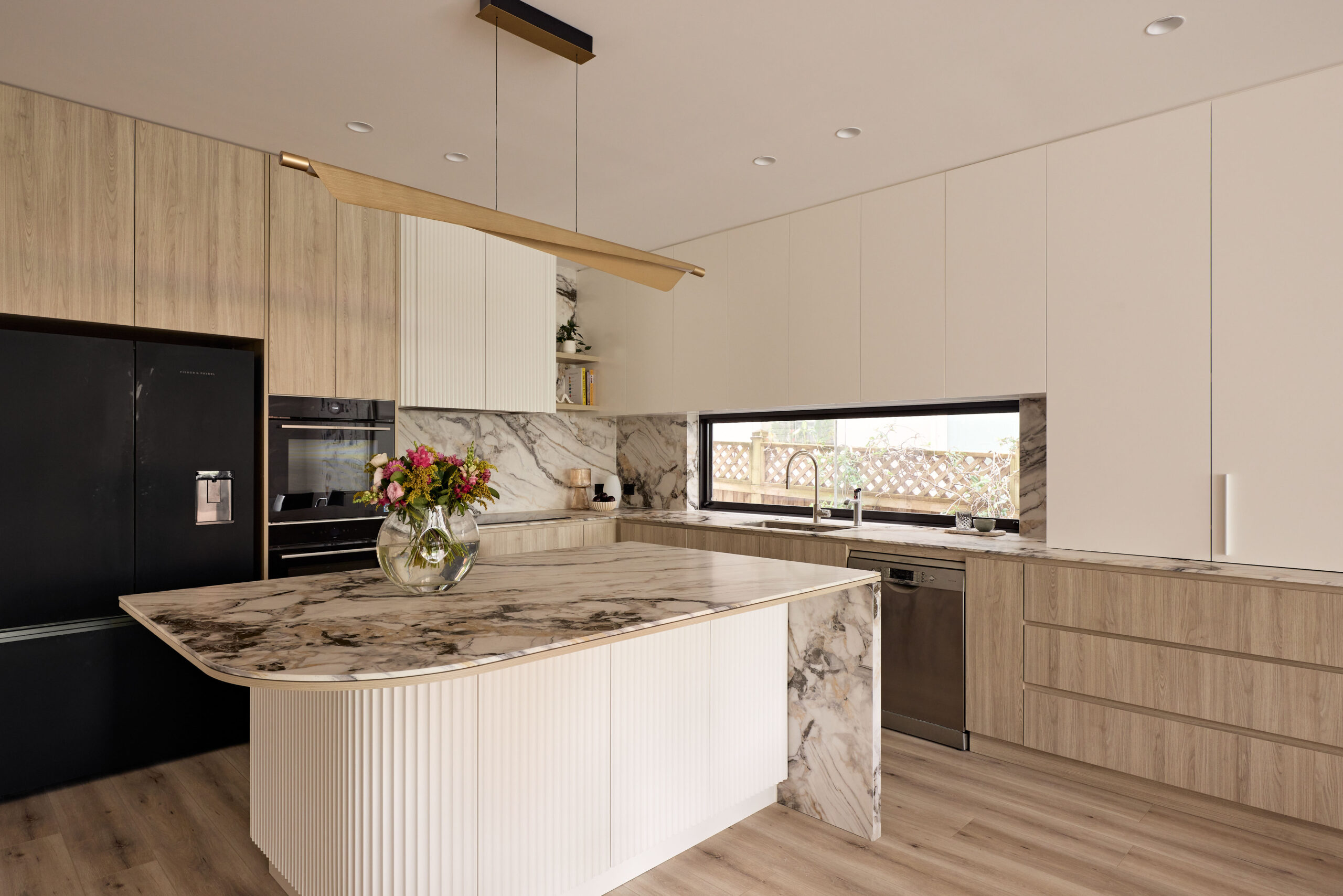 Modern kitchen with marble countertops, light wood cabinetry, a black fridge, and a long wood pendant light above the island.