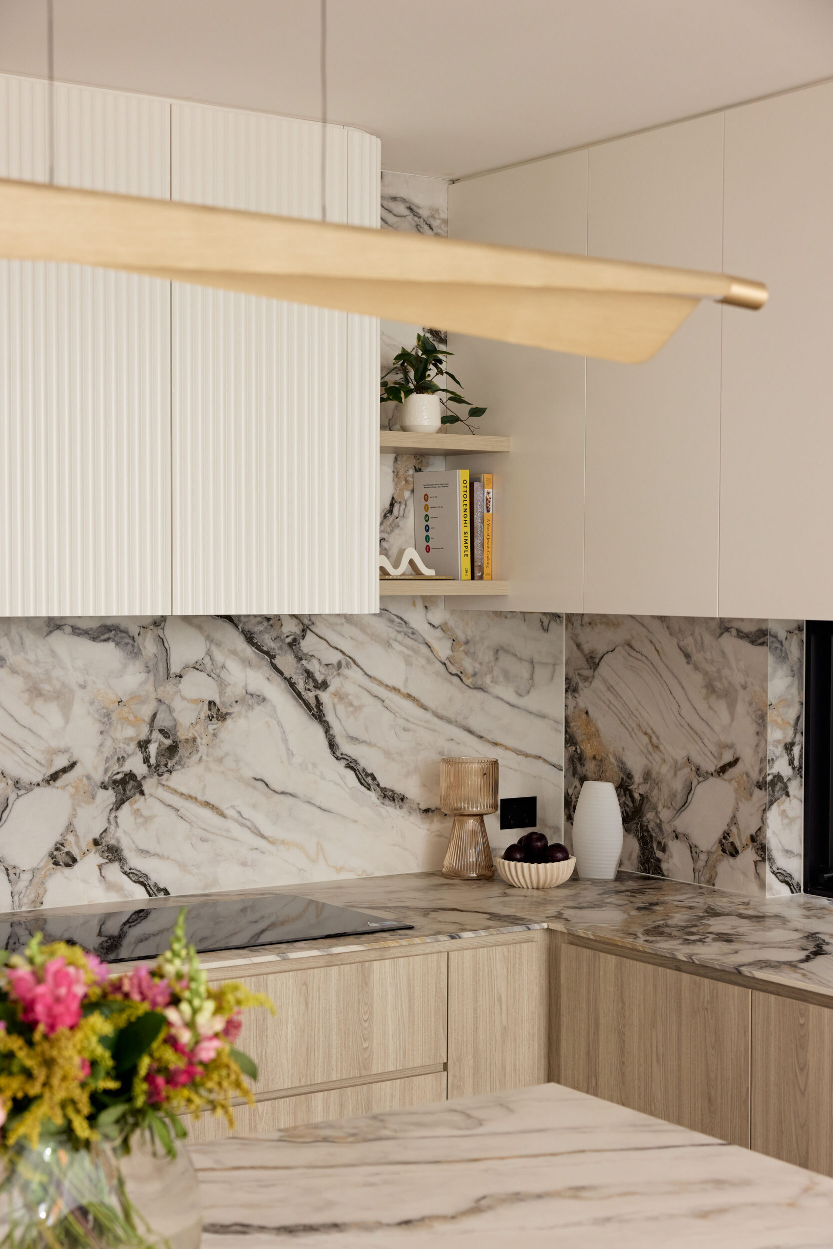 Modern kitchen with white paneled cabinets, marble backsplash and countertop, a small shelf with a plant and books, and a long wooden pendant light across the scene.