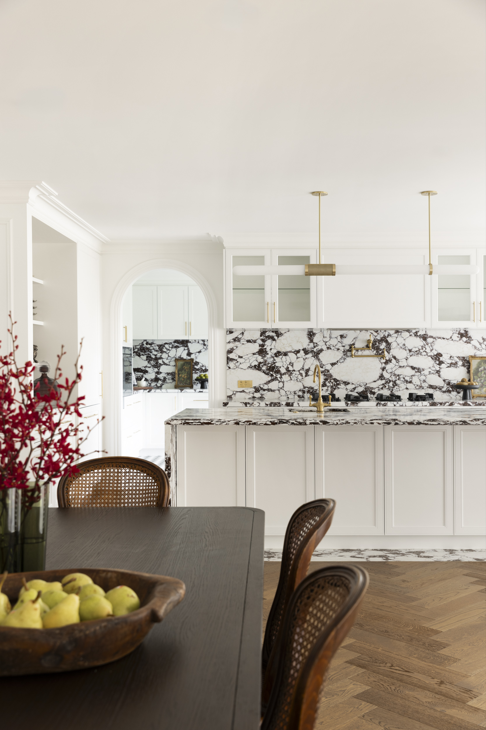 Bright white kitchen with a marble black-and-white island, gold fixtures, and a dark dining table in the foreground with a fruit bowl and wicker chairs nearby.