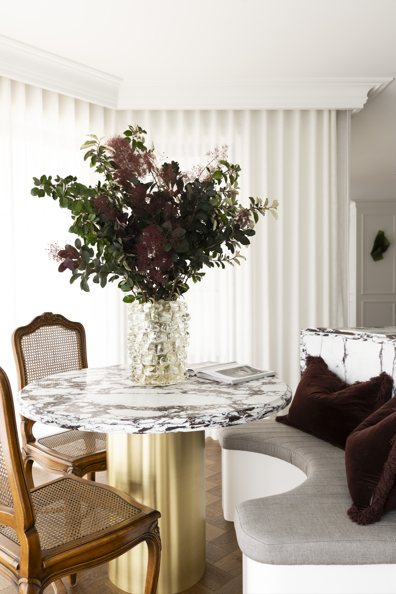 Elegant dining nook with a round marble table, gold pedestal base, and a tall vase of greenery and burgundy flowers, next to cane chairs and a light sofa with pillows.