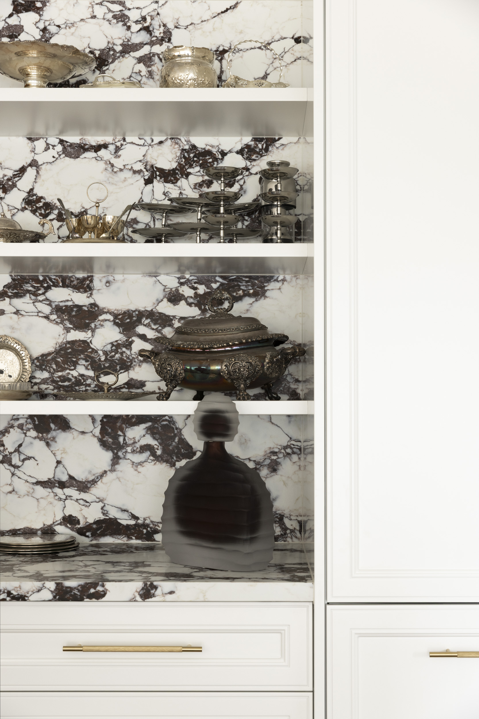Open cabinet with white shelves and black-and-white marble back, displaying silver servingware and a large ornate tureen.