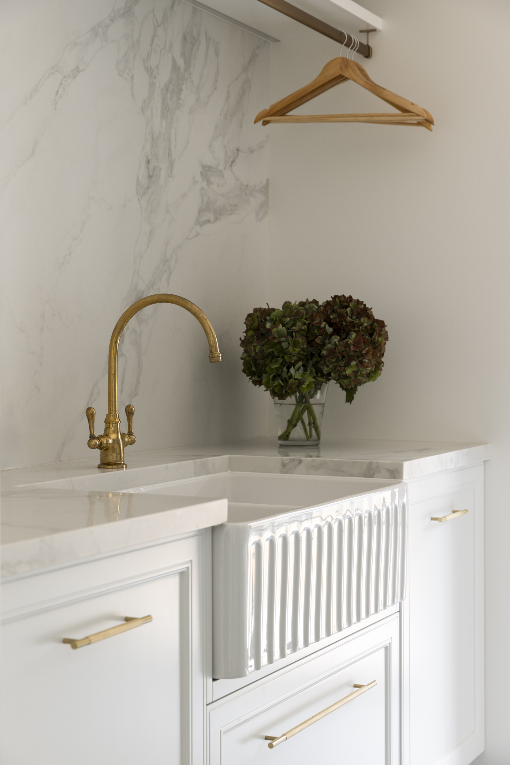 White kitchen scene with marble backsplash, a brass faucet, a vase of green hydrangeas on the counter, and wooden hangers hanging above.