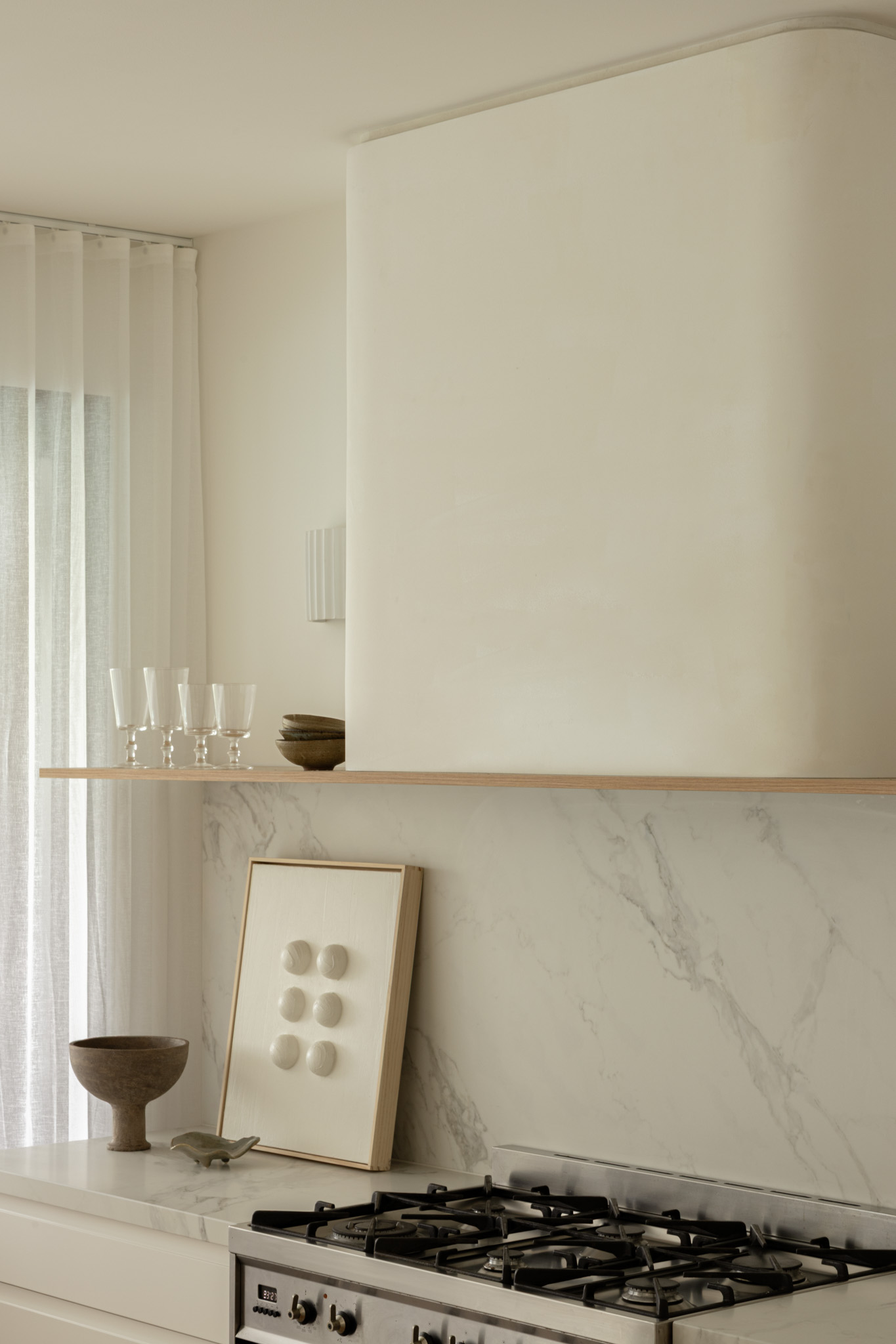 Minimalist kitchen scene with a marble backsplash, gas stove, and a wooden shelf holding a frame with white knobs and a bowl on the counter.