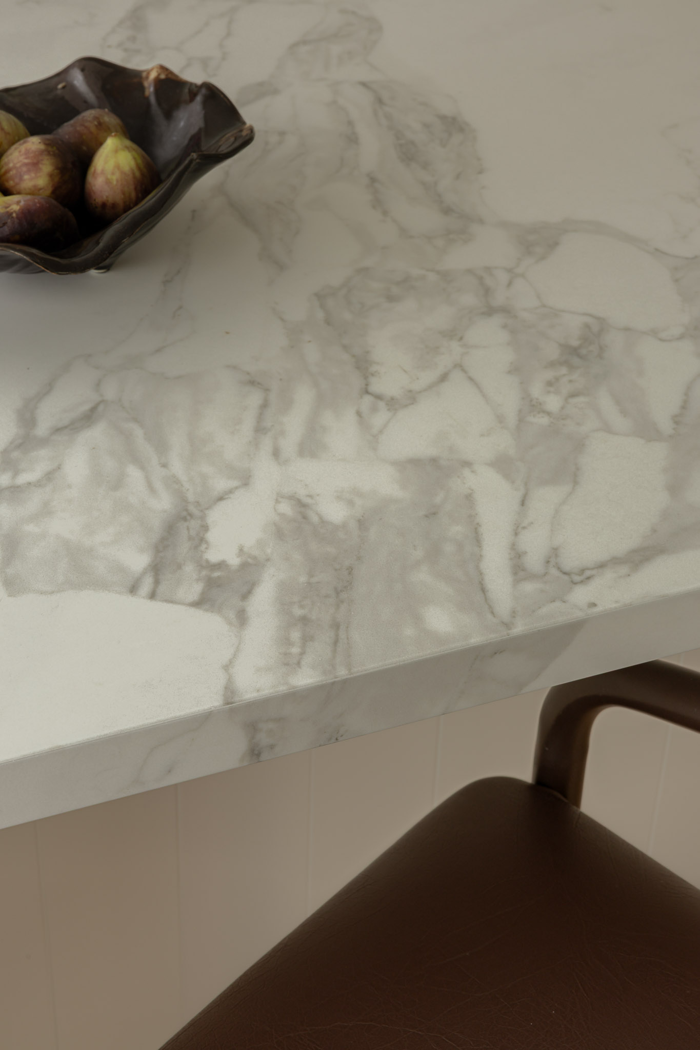 Dark brown ceramic bowl filled with figs sits on a white marble kitchen counter, with part of a brown chair visible in the foreground.