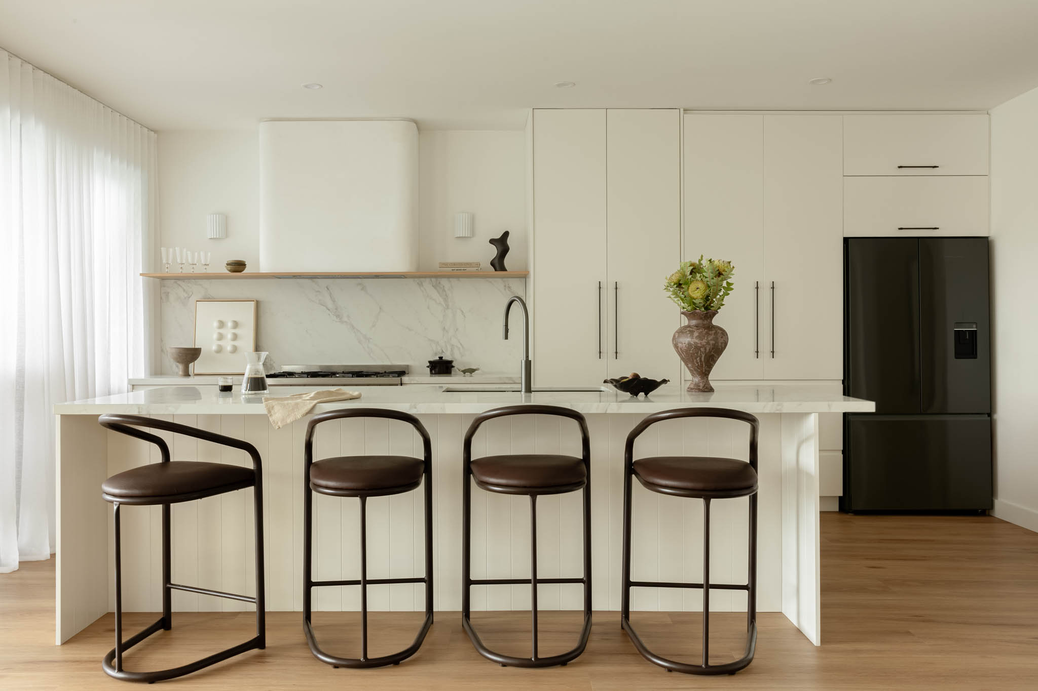 Bright modern kitchen with white cabinets, marble island, and four brown metal bar stools along a large island