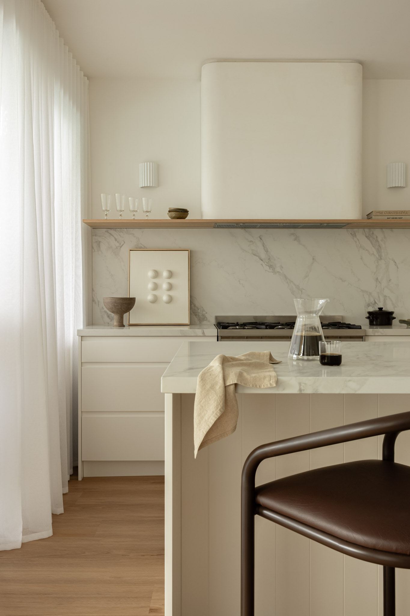 Minimalist kitchen with white marble island, pale wood accents, and sheer white curtains on left