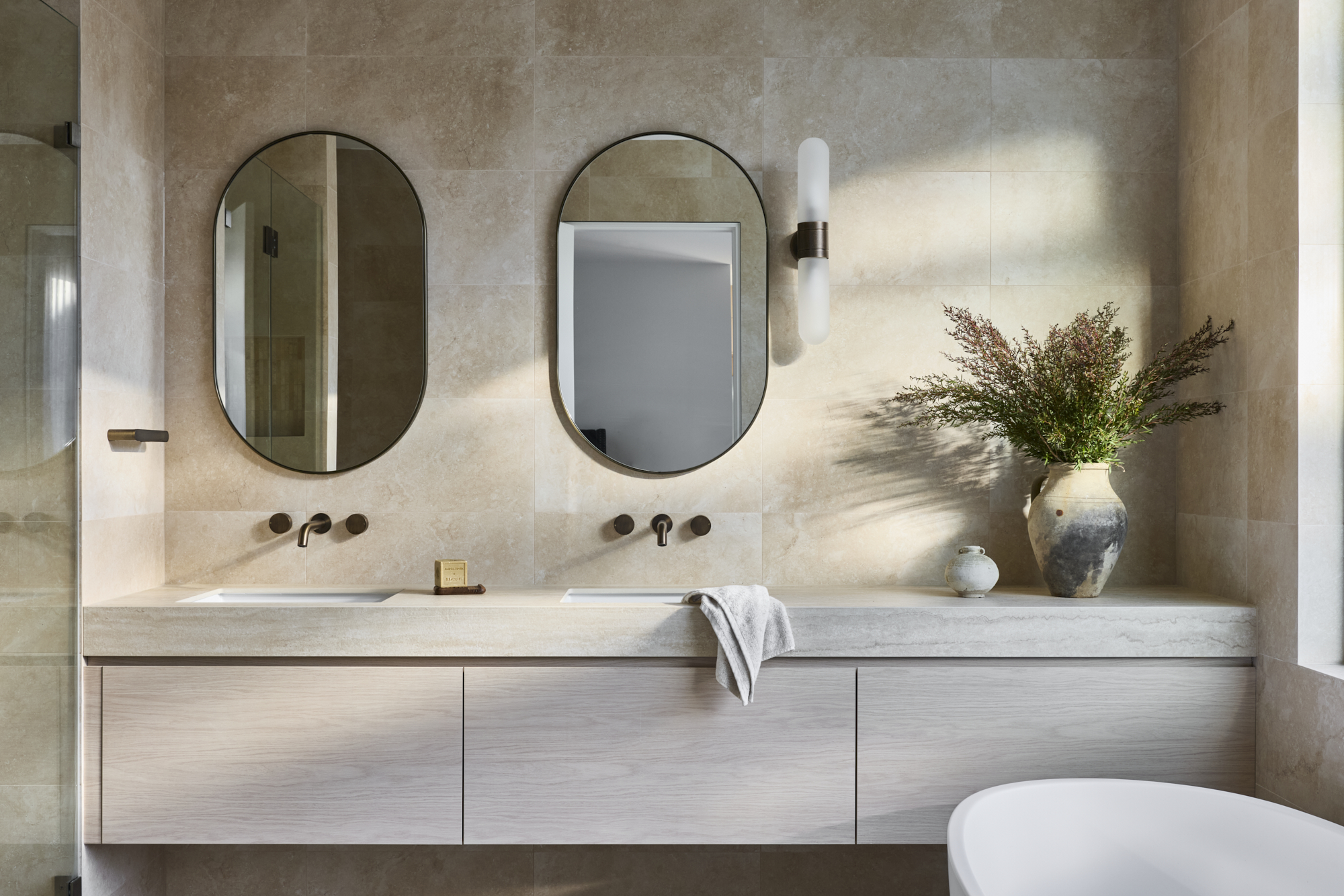 Contemporary bathroom vanity with two black oval mirrors, beige tile walls, and a tall vase of greenery on the right.