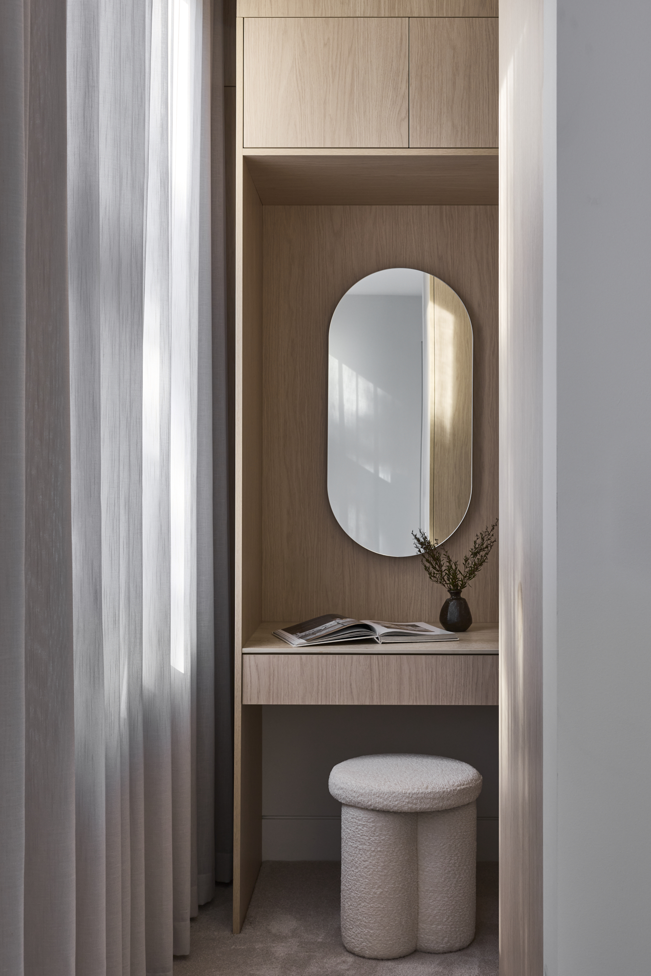 Wooden built-in vanity area with an oval mirror, magazines on the desk, and a small vase, framed by soft curtain panels on the left.