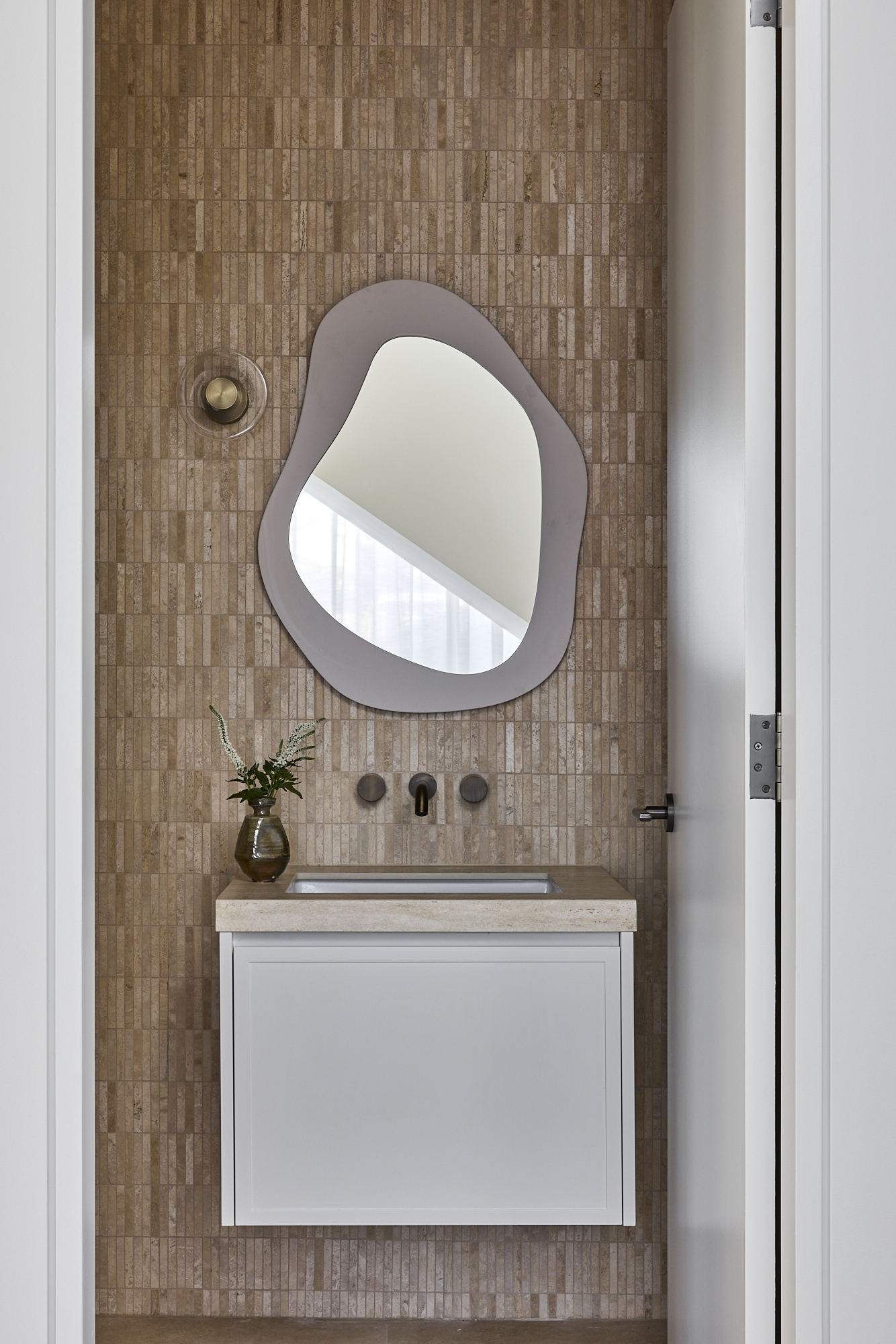 Bathroom vanity with a white cabinet, beige tiled wall, and an irregular rounded mirror above the sink; a brown vase with greenery on the counter.
