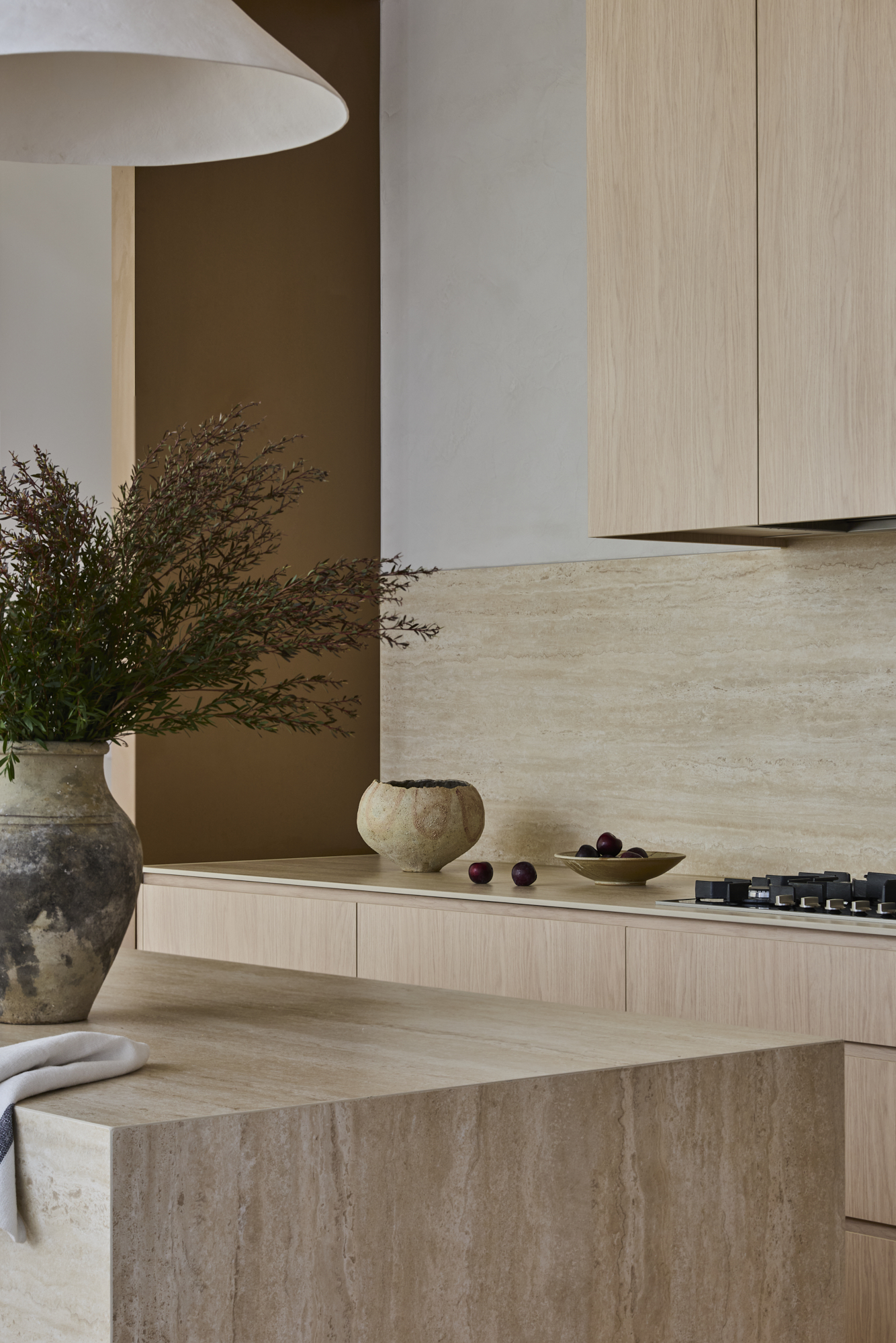 Modern kitchen scene with light wood cabinets, beige stone countertops, and a large ceramic vase with greenery on the left.
