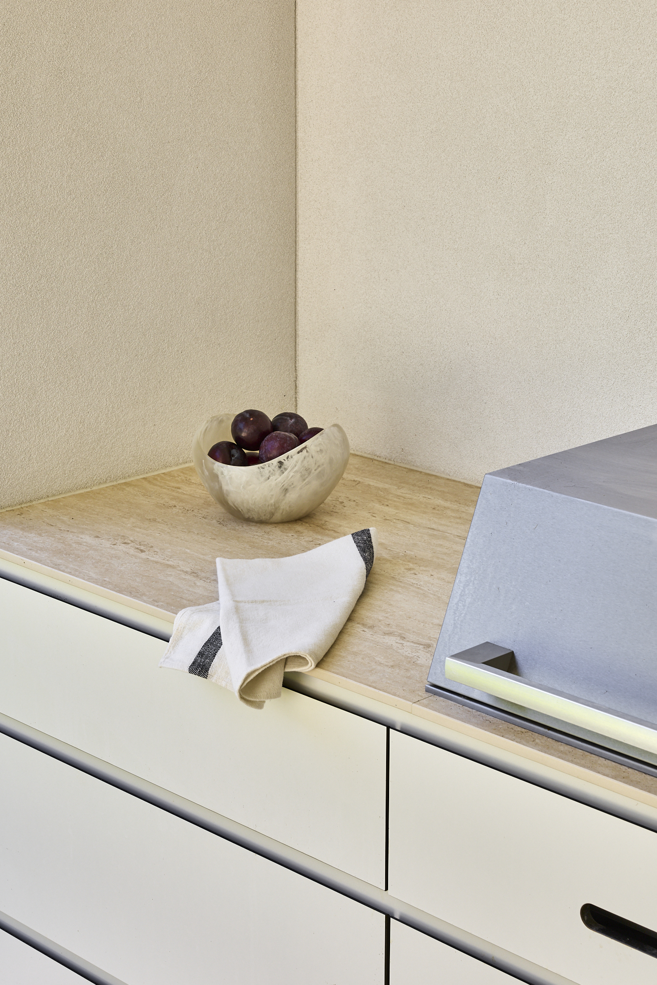 White marble bowl filled with dark plums on a light wooden kitchen counter beside a stainless range hood.