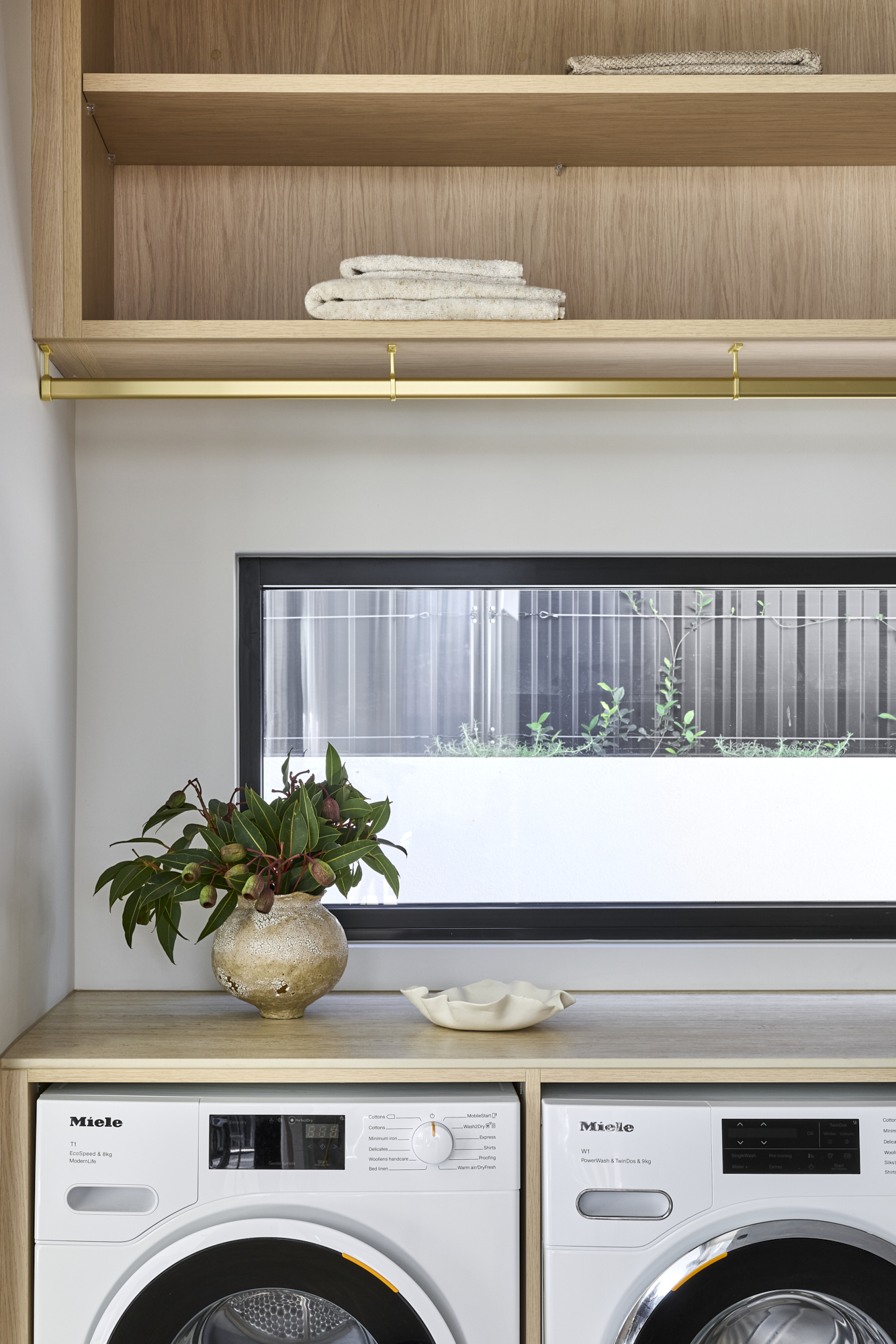 Laundry alcove with two front-loading washers under a wooden counter, a brass hanging rail above, and a plant on the counter.