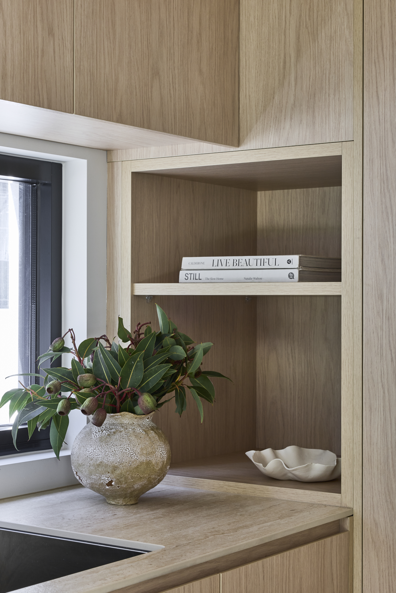 Wooden cabinet shelf with a ceramic vase of greenery and two stacked books (Live Beautiful, Still).