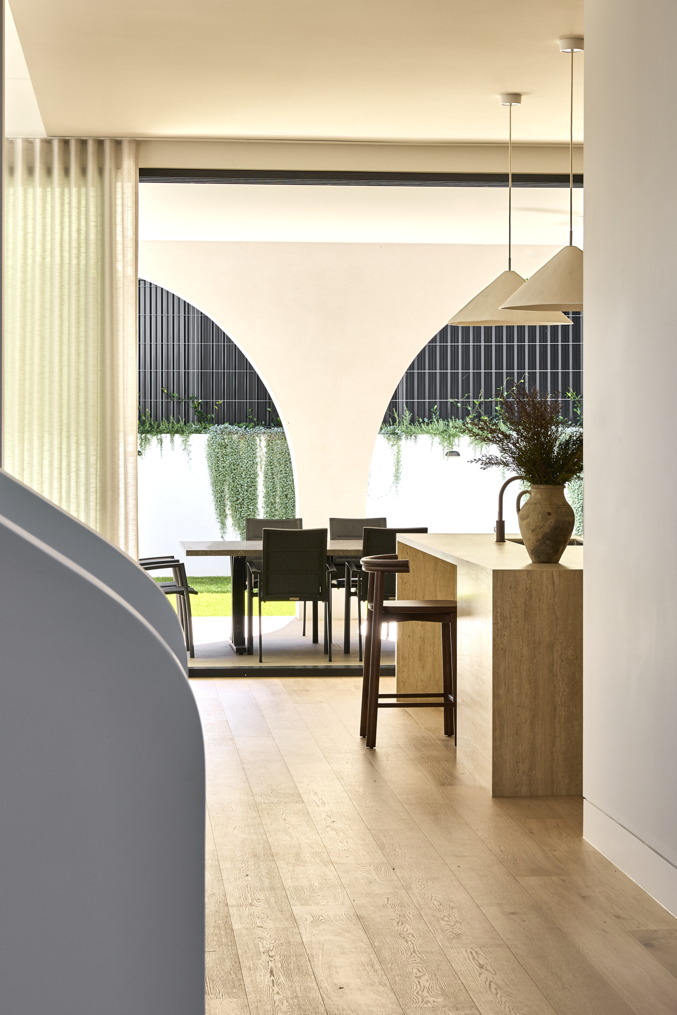 Bright modern dining area with a long table and stools, seen through large curved archways to a garden outside.
