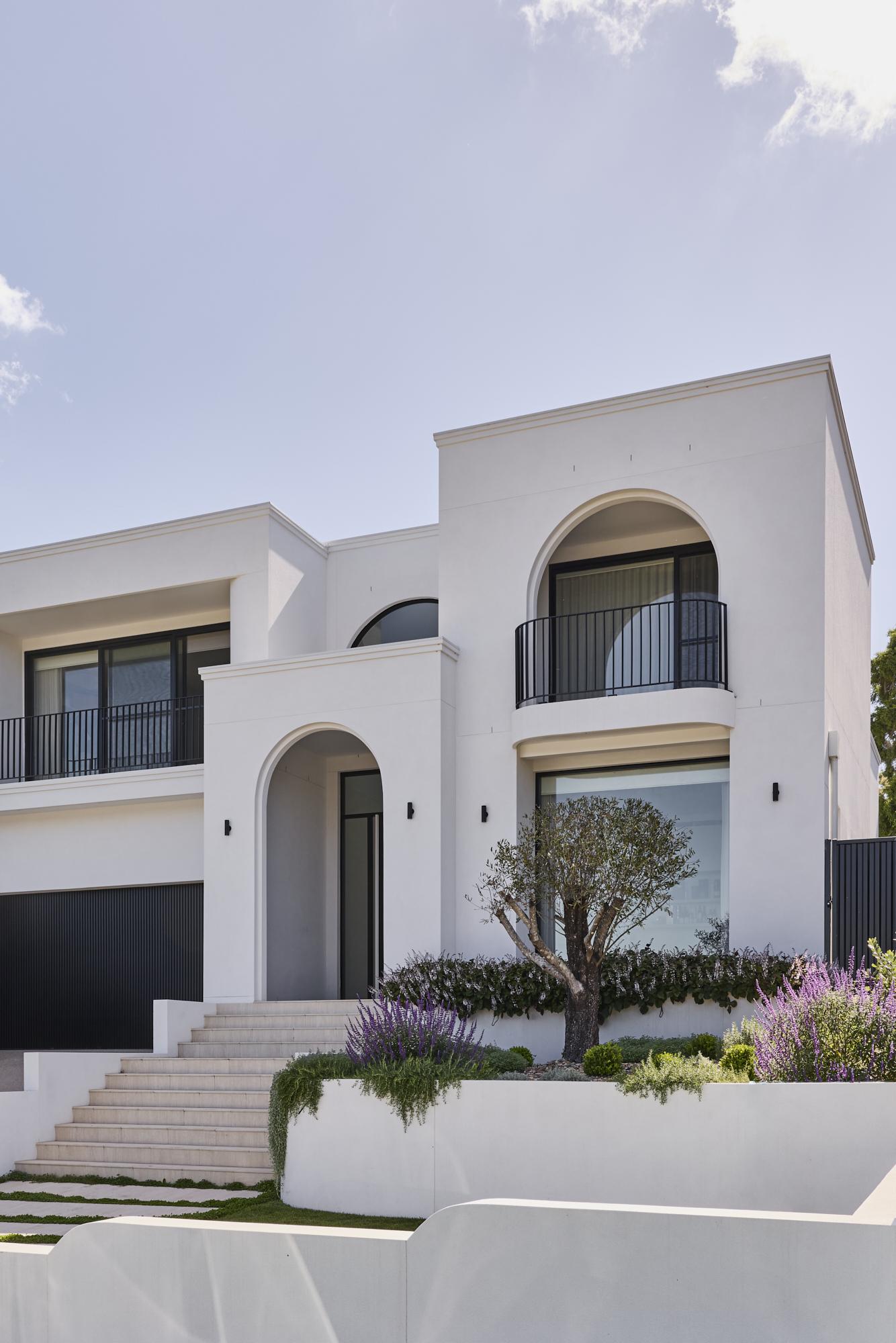 White modern building with arched balcony and black railings, set against a clear blue sky and a landscaped front yard with purple flowers.