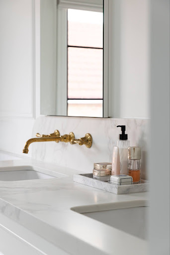 White marble bathroom counter with a gold wall-mounted faucet, mirror, and a tray of toiletries by the sink.