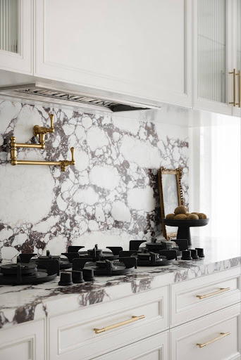 White kitchen with marble backsplash and countertop, brass faucet, and a six-burner gas stove; framed mirror and a fruit bowl on the counter.