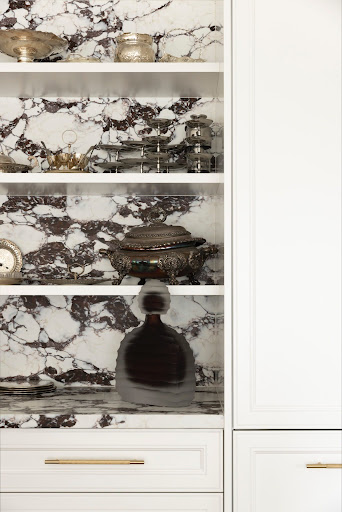 Shelving unit with white cabinetry and dark marble backdrop, holding decorative items including gold bowls, a metal rack, and a dark glass decanter.