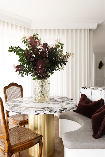 Round marble dining table with a glass vase of burgundy and green flowers on a gold pedestal base, surrounded by wooden chairs in a bright living room.