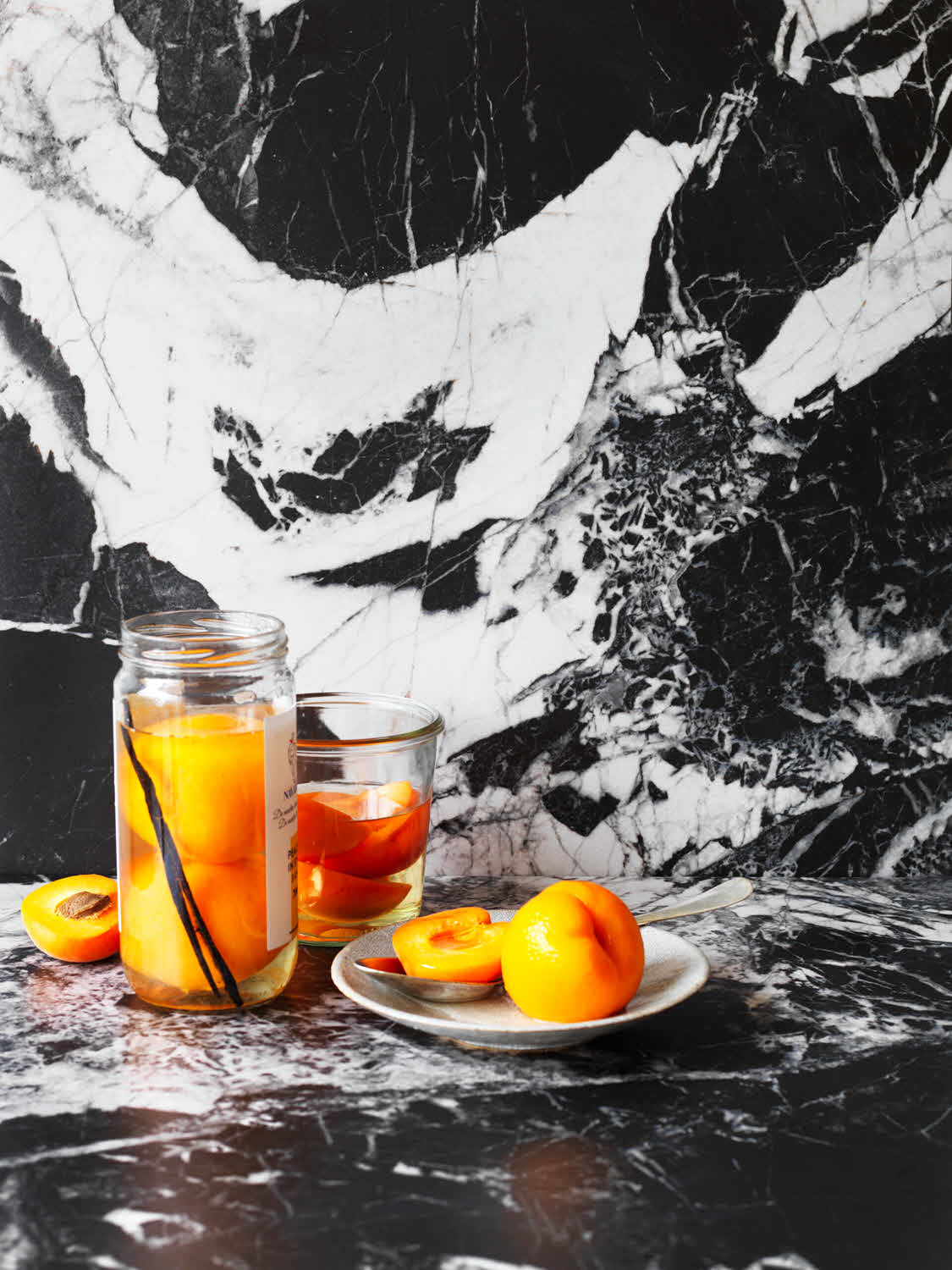 Two glass jars of orange juice with orange slices inside, set on a white-and-black marble surface, with halved apricots nearby and a vanilla bean in the left jar.