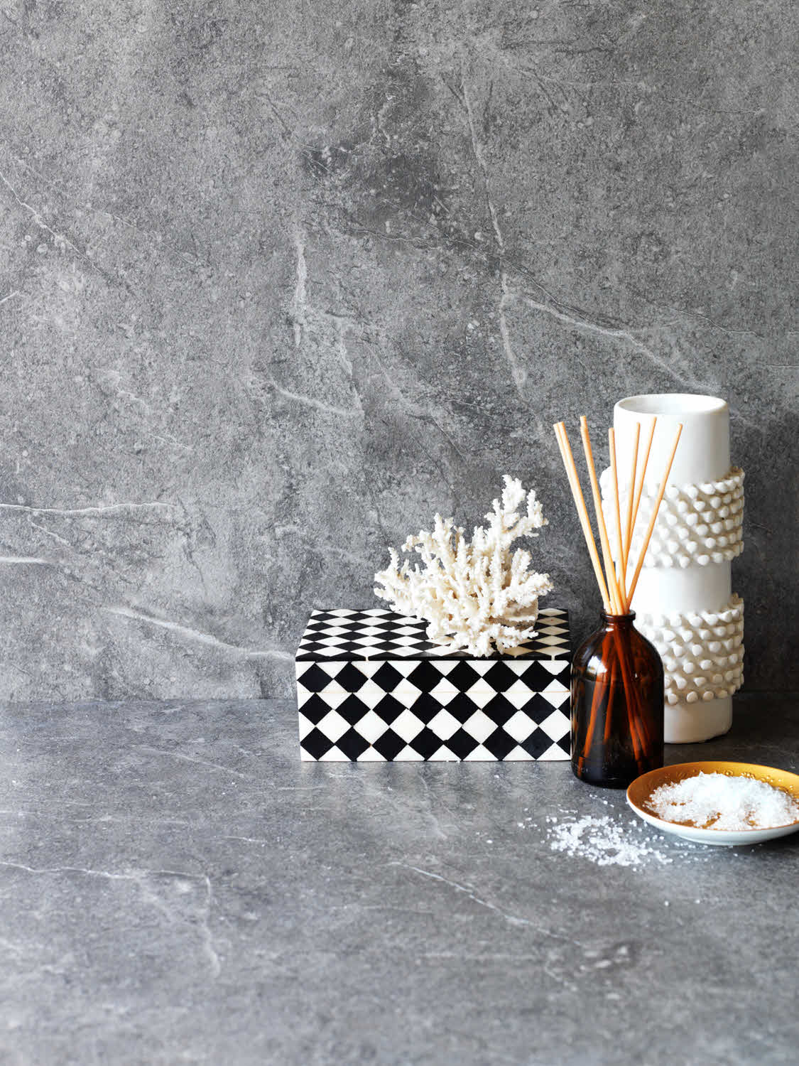 Still life arrangement on a gray stone surface: a black-and-white geometric box, white coral, an amber diffuser bottle with reeds, and a textured white vase at right show minimal decor.