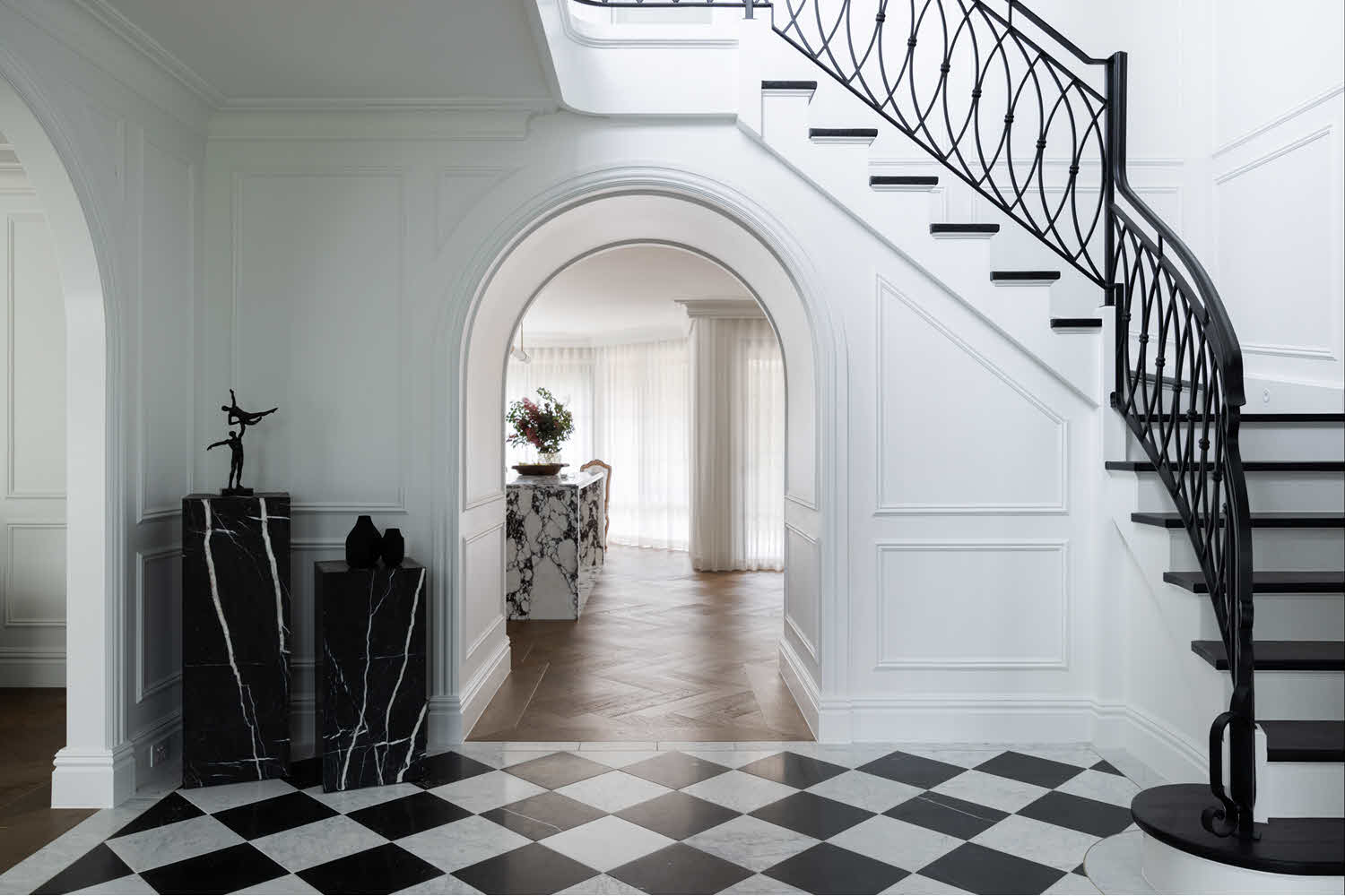 Elegant foyer with a black-and-white checkered floor, a curved archway, marble pedestals and sculpture on the left, and a sweeping black wrought-iron staircase on the right.