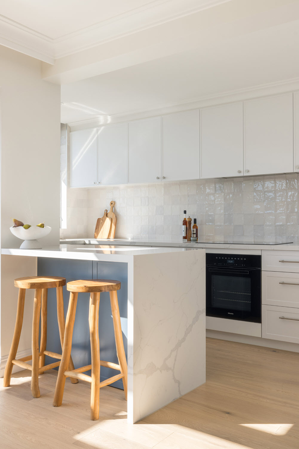 Bright white modern kitchen with a marble island, two wooden bar stools, and a tiled backsplash under soft natural light.