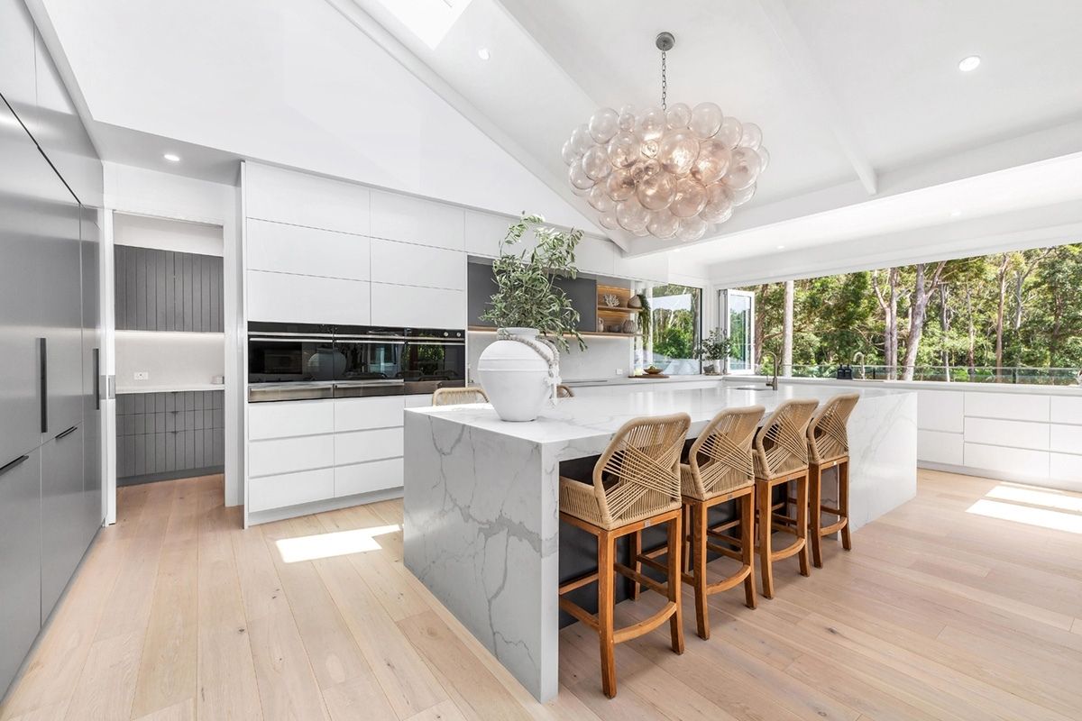 Bright modern kitchen with a marble island, white cabinets, and woven wooden bar stools overlooking greenery outside.
