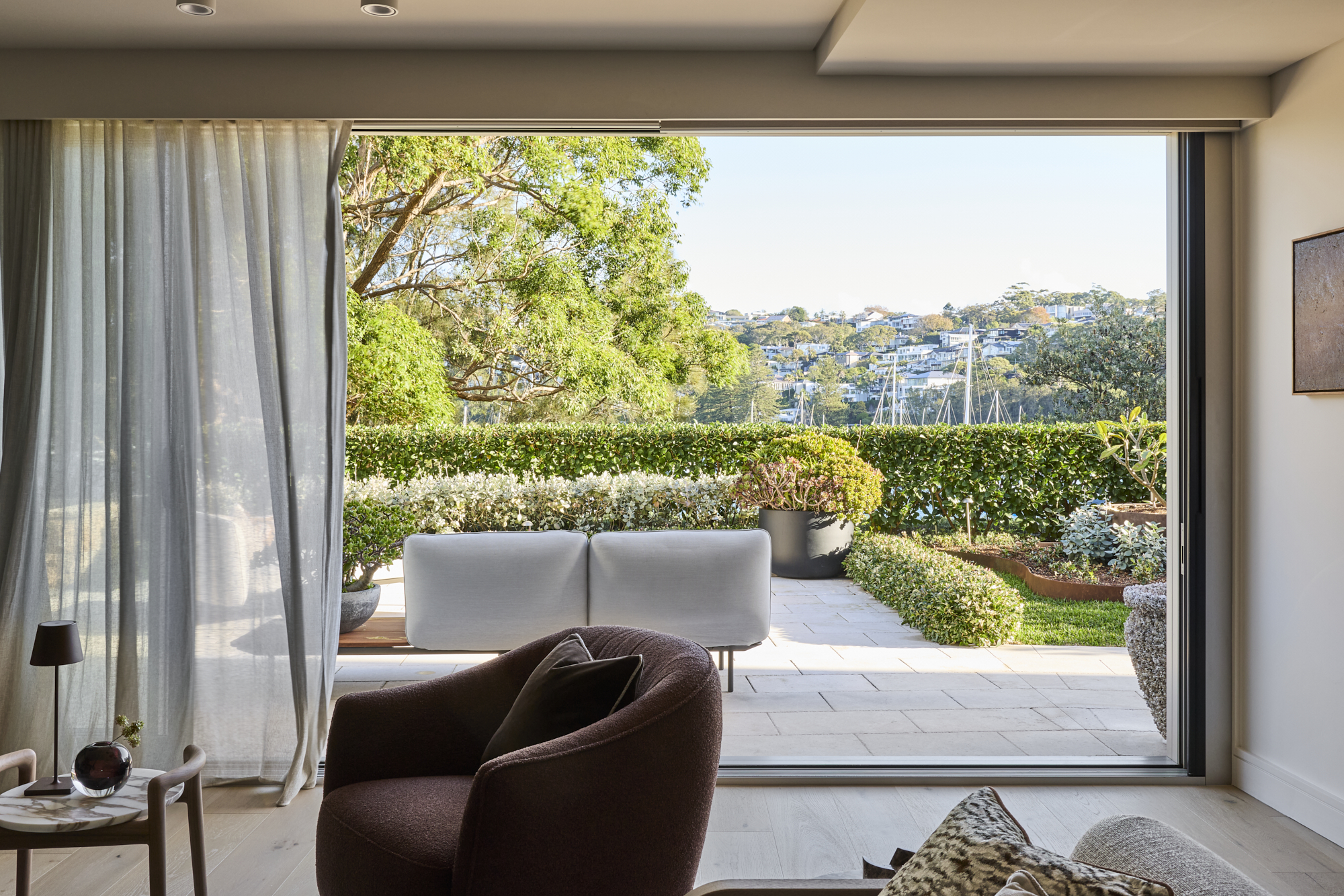 Living room with a wide open sliding glass door revealing a sunny garden with trees, hedges, and patio seating.