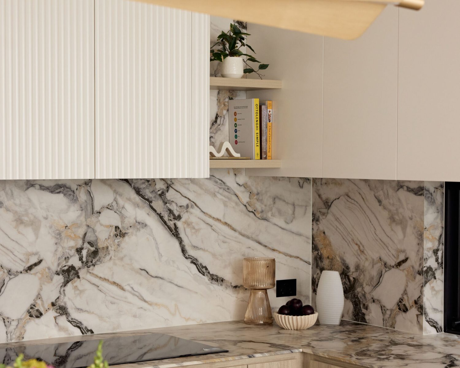 Modern kitchen with white paneled cabinets, marble backsplash and countertop, a small shelf with a plant and books, and a long wooden pendant light across the scene.