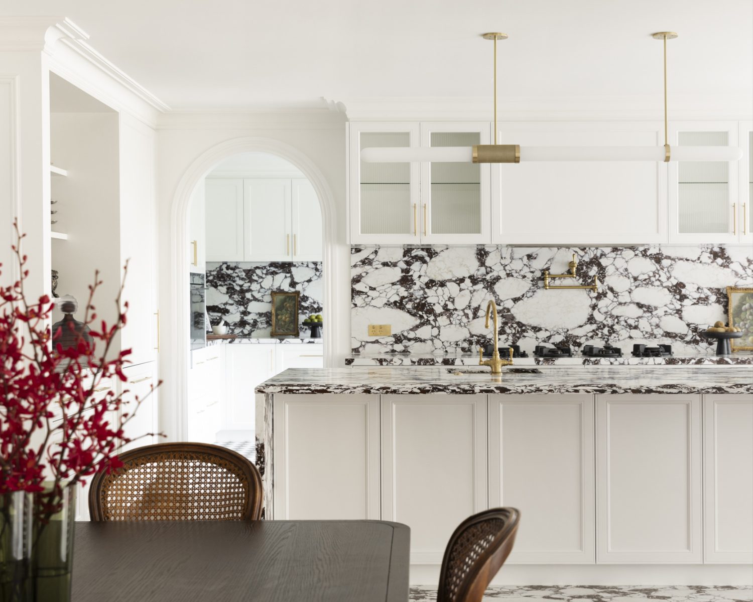 Bright white kitchen with a marble black-and-white island, gold fixtures, and a dark dining table in the foreground with a fruit bowl and wicker chairs nearby.