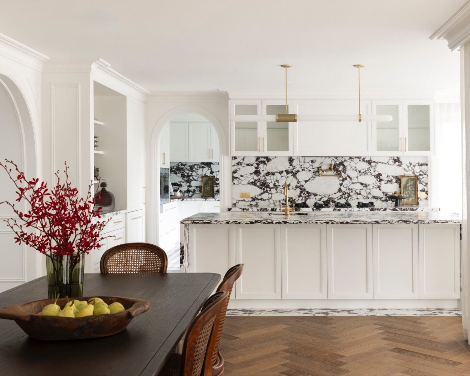 Bright white kitchen with marble backsplash and island, gold hardware, and a dark dining table in the foreground with red branches in a vase.